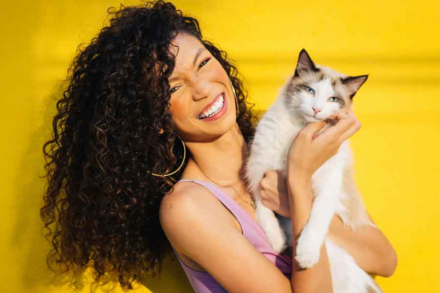 A woman with brown curly hair holds a white cat with brown ears and smiles at the viewer from a bright yellow background.