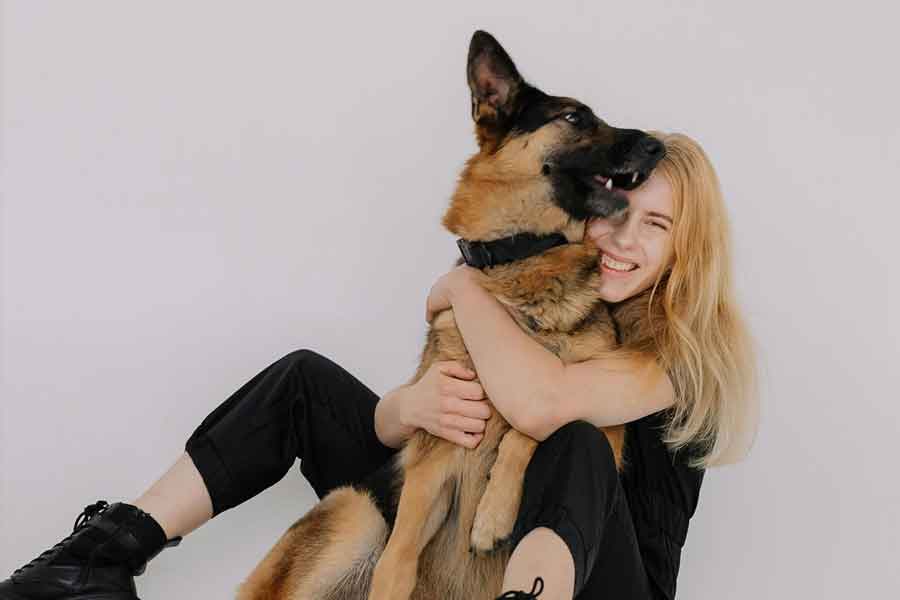 A woman with blond hair wearing black sits behind a german shepherd and hugs it tightly.