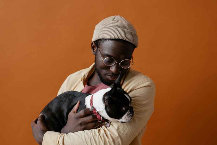 A man with dark brown skin and round glasses wearing tan hugs a black and white Boston terrier dog and smiles down at it from an orange background.